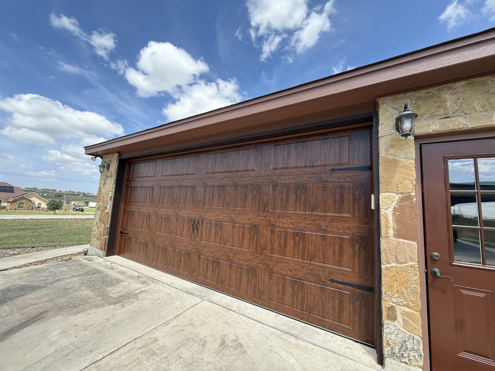 Clopay 18x8 walnut wood-grain insulated garage door with black decorative hardware on a stone home in Buda, TX